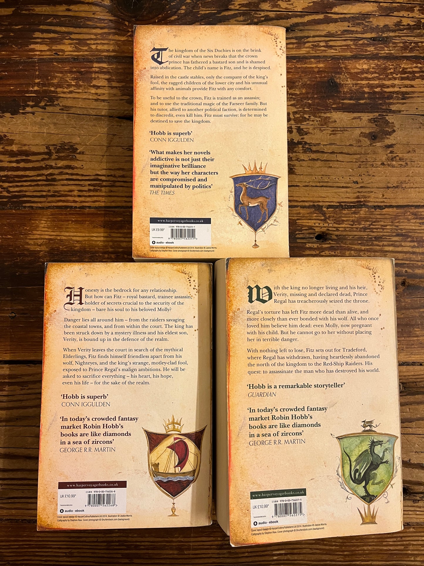 Back cover of three books titled 'Assassin's Quest', 'Royal Assassin', and 'Assassin's Apprentice' by Robin Hobb on a wooden surface.
