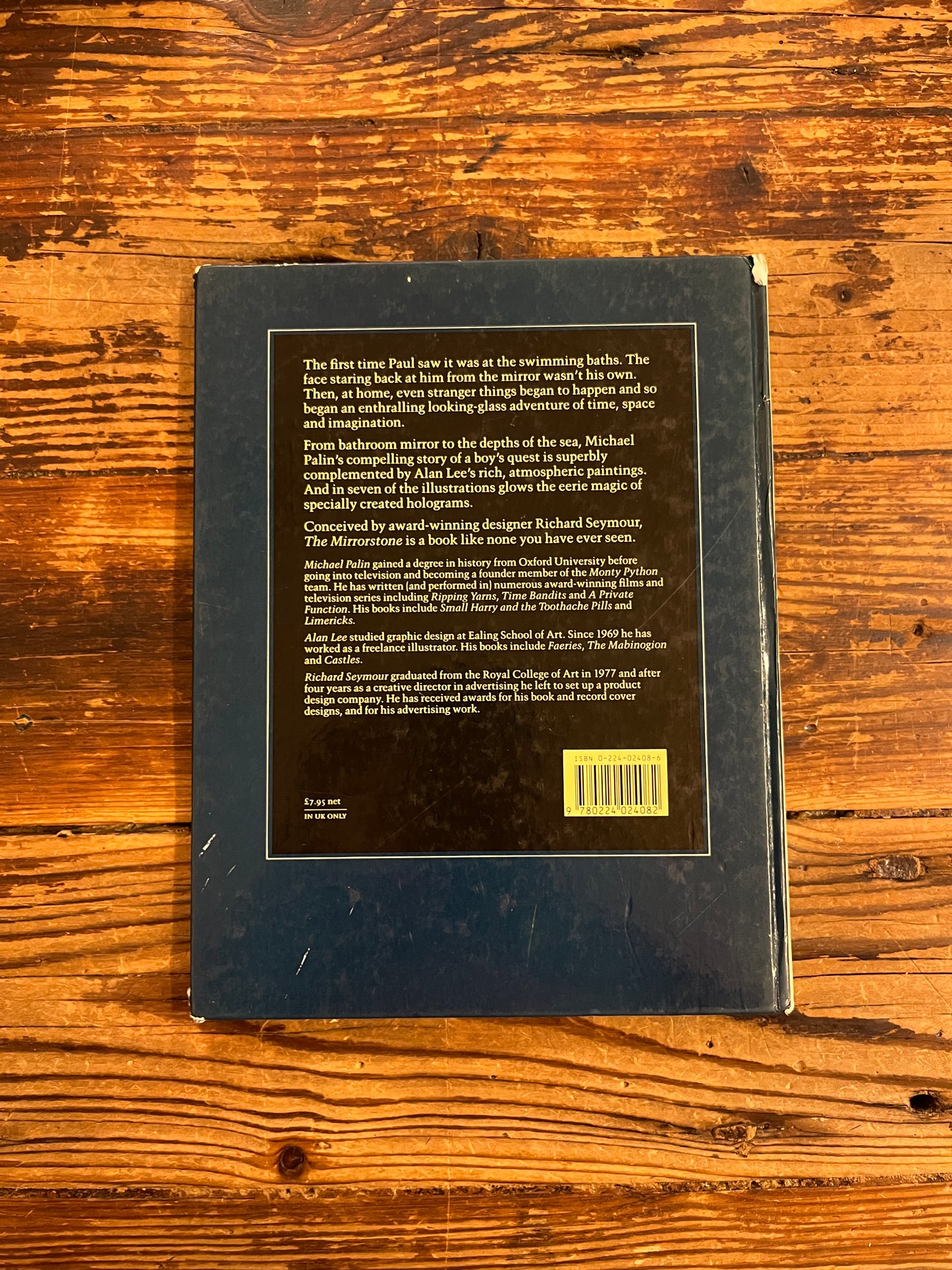 Back cover of book titled 'The Mirrorstone' on a wooden surface showing a damaged corner