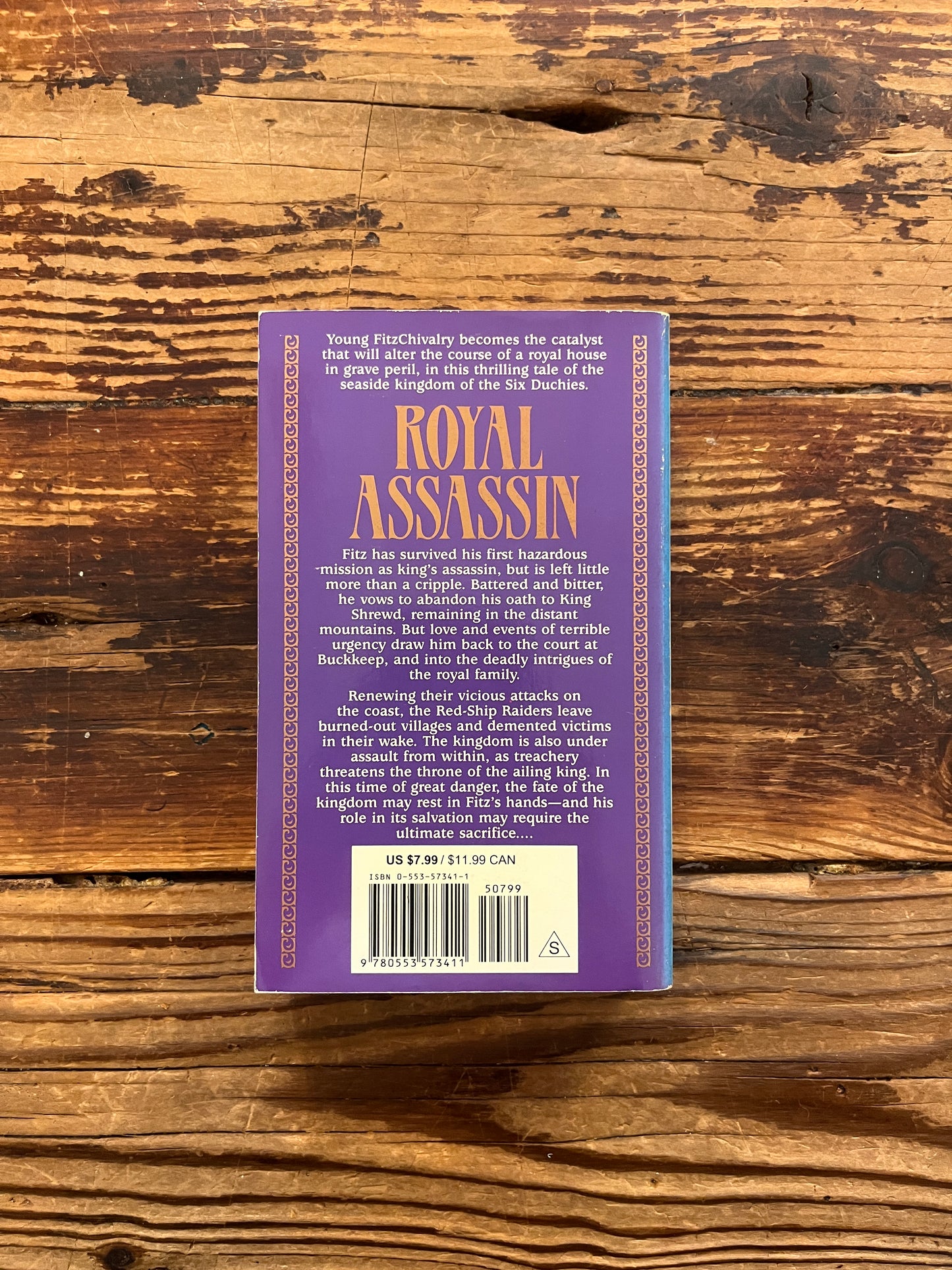 Back cover of a book titled 'Royal Assassin' by Robin Hobb on a wooden surface