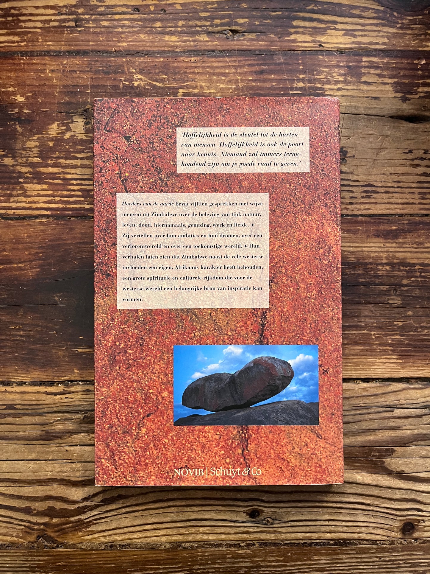 Achterkant van boek met de titel 'Hoeders van de Aarde' door Chenjerai Hove en Ilija Trojanow op een houten achtergrond