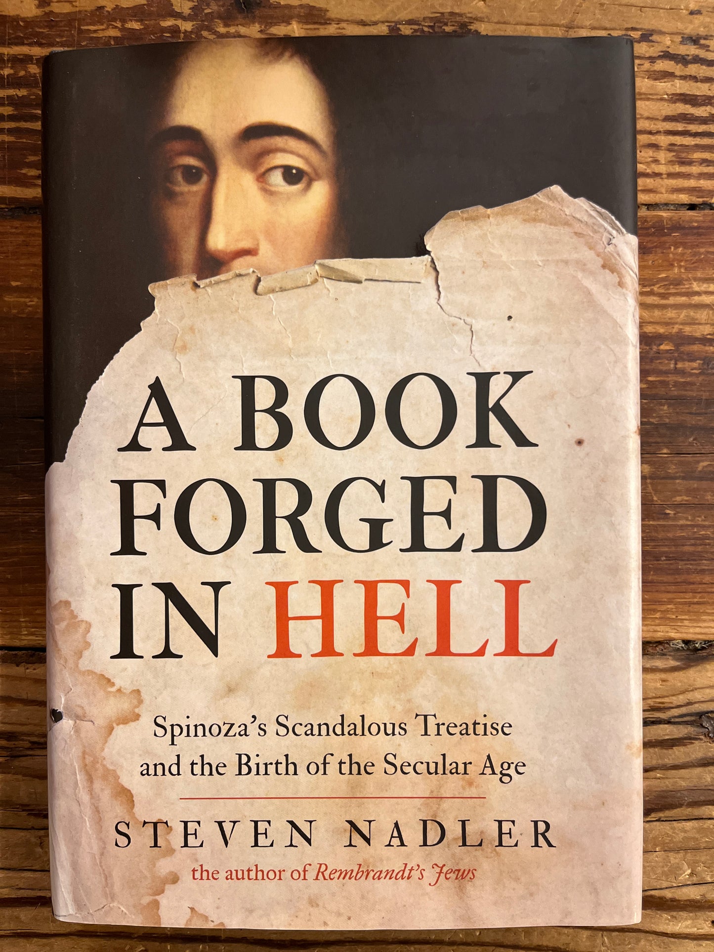 front cover of book titled 'A Book Forged in Hell' by Steven Nadler on a wooden surface