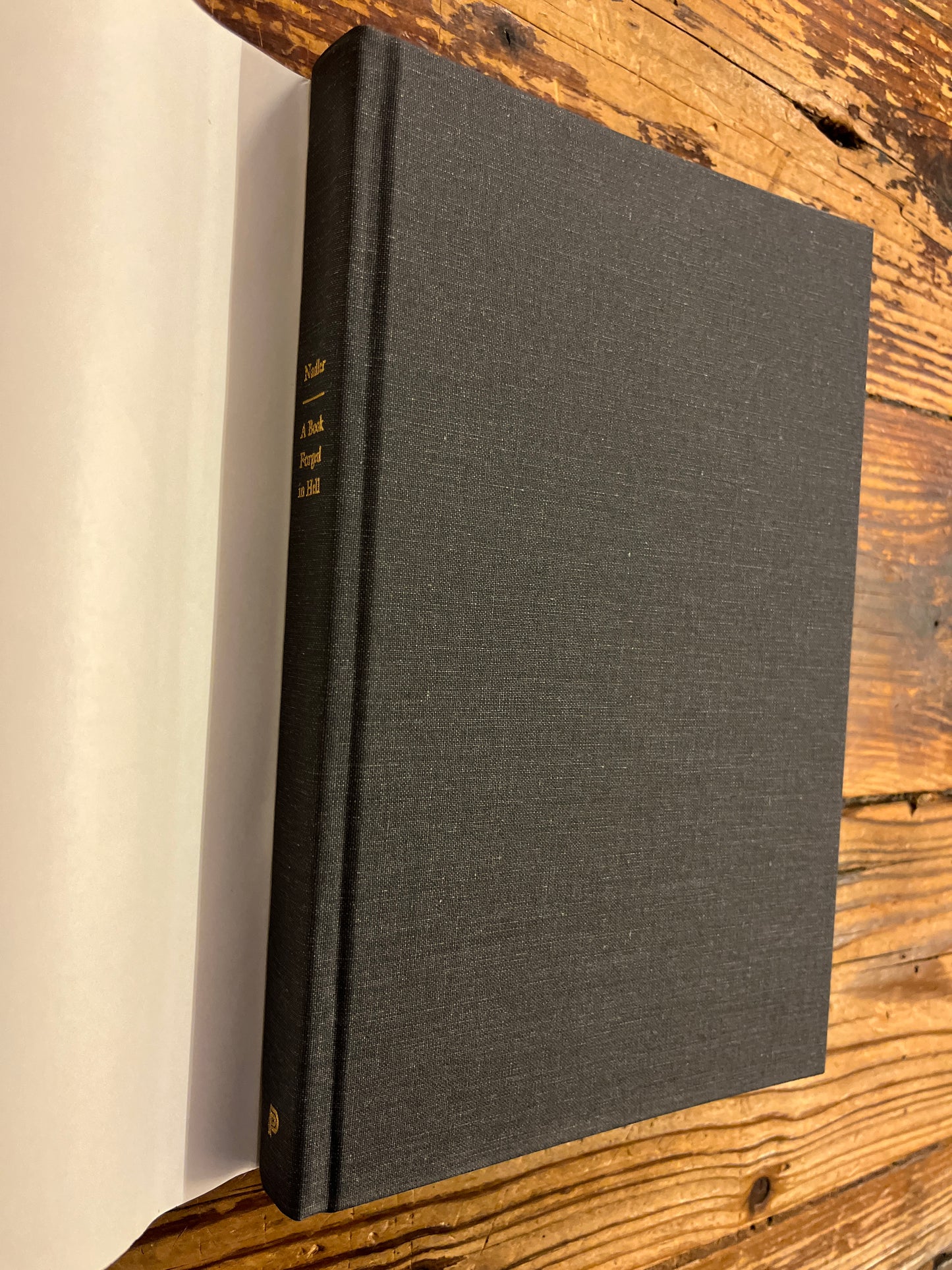 Black bound book titled 'A Book Forged in Hell' by Steven Nadler on a wooden surface on a wooden surface