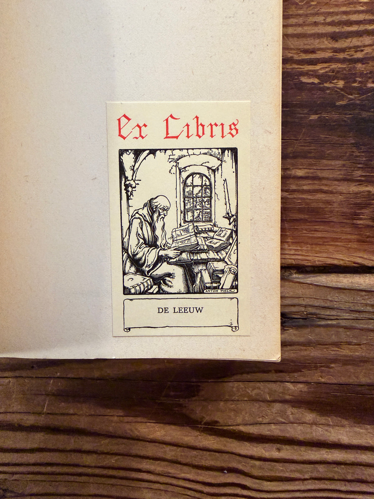 Eerste pagina met ex-libris sticker van boek met de titel 'Uitgenodigd, l'invitée' door Simone de Beauvoir op een houten achtergrond
