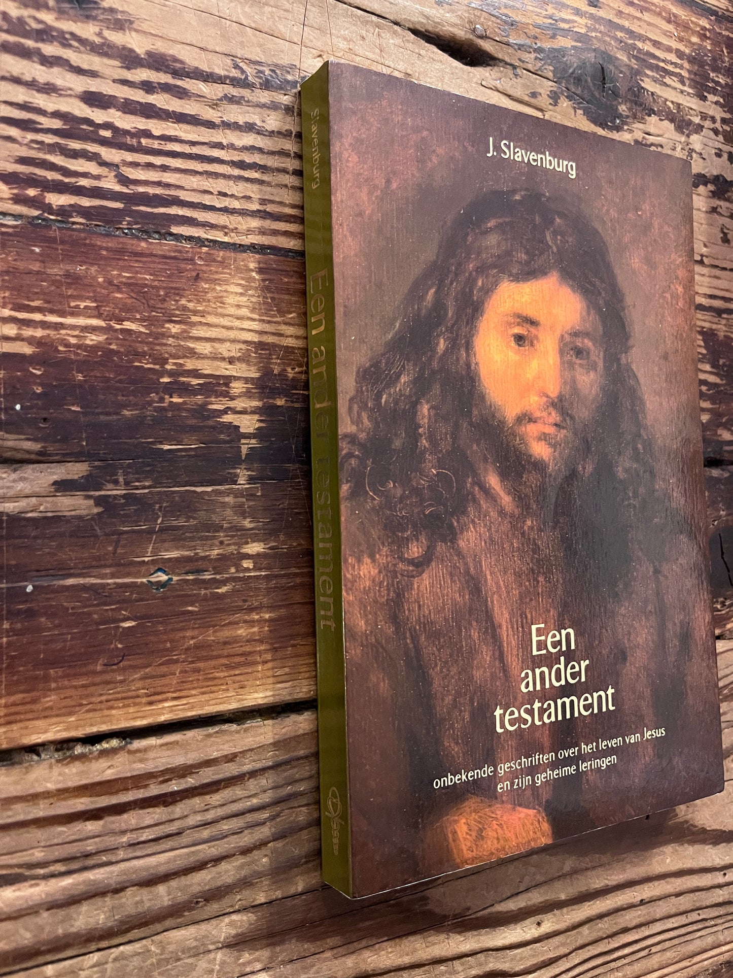 Voorkant en rug van boek met de titel 'Een ander testament' door J. Slavenburg op een houten achtergrond 