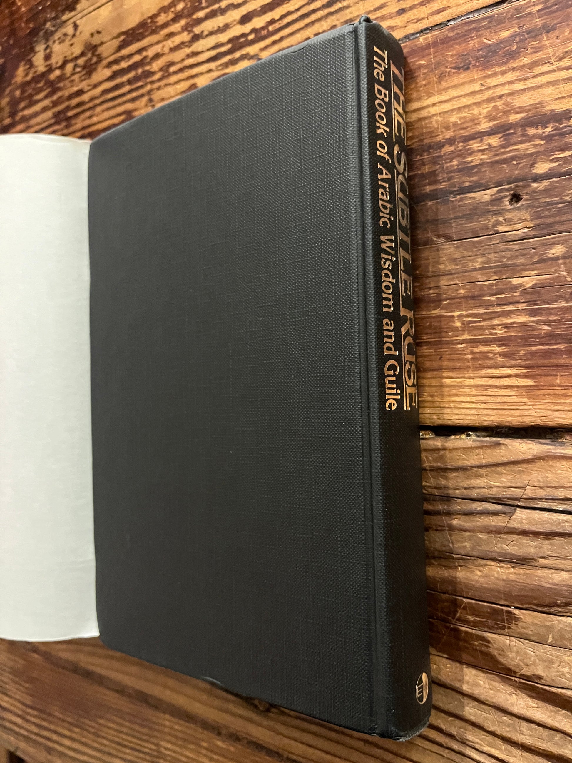 This image features the spine and back cover without dust jacket of a hardcover book titled "The Subtle Ruse, The Book of Arabic Wisdom and Guile," translated by Rene R. Khawam on a wooden surface