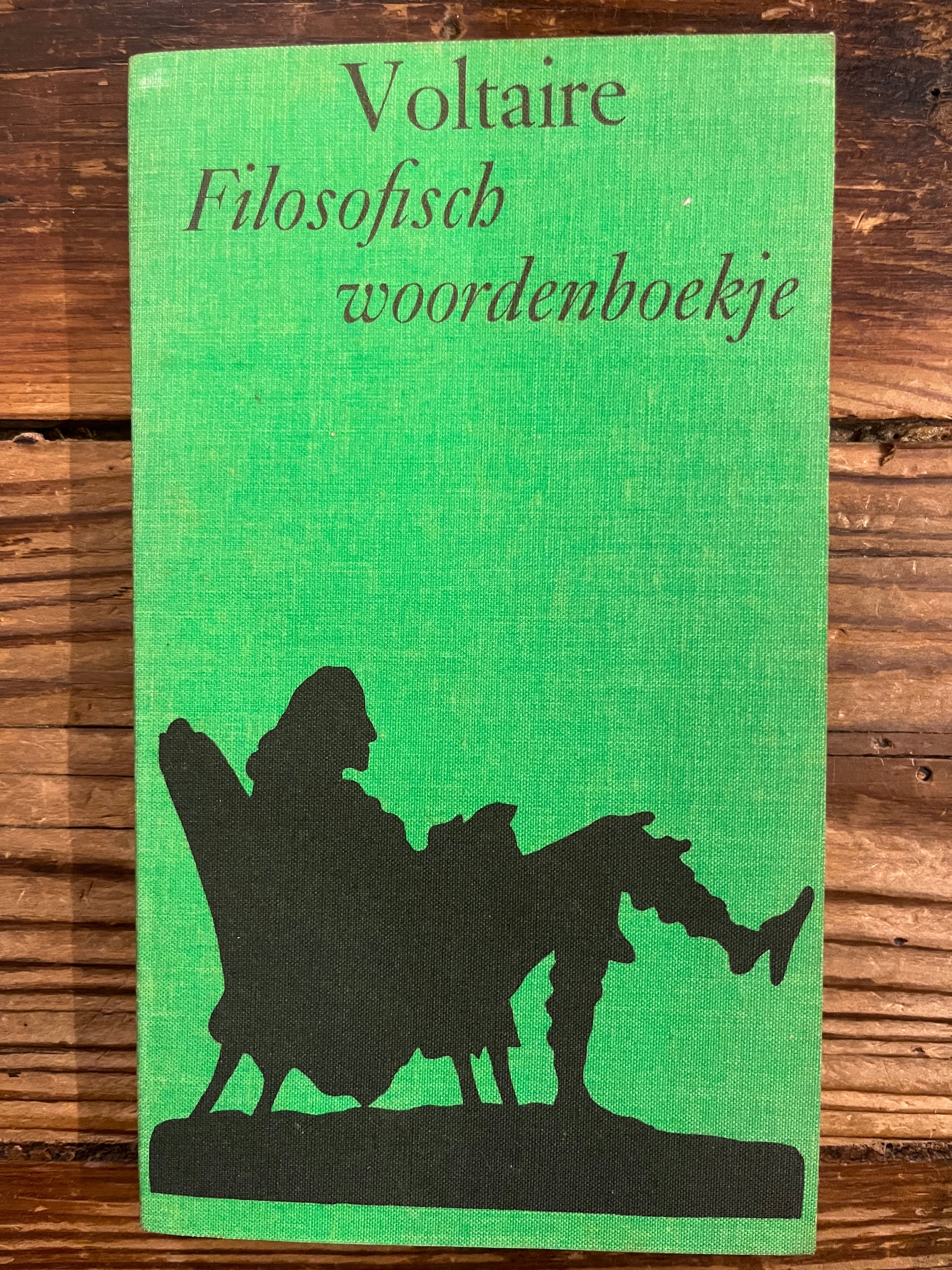 Voorkant van boek met de titel 'Filosofisch woordenboekje' door Voltaire op een houten achtergrond 