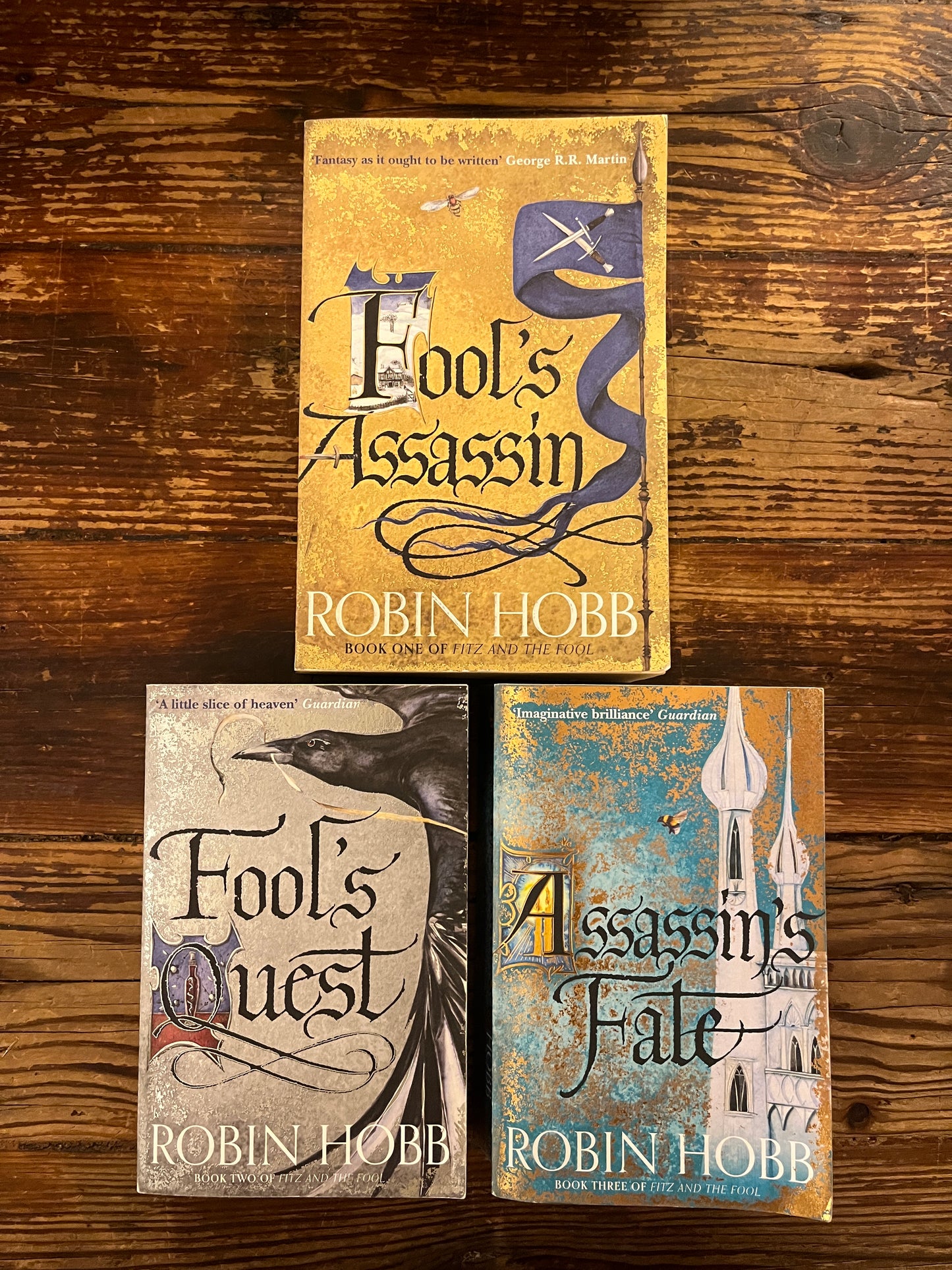 Three books titled 'Fool's Assassin', 'Fool's Fate', and 'Fool's Quest' by Robin Hobb on a wooden surface.