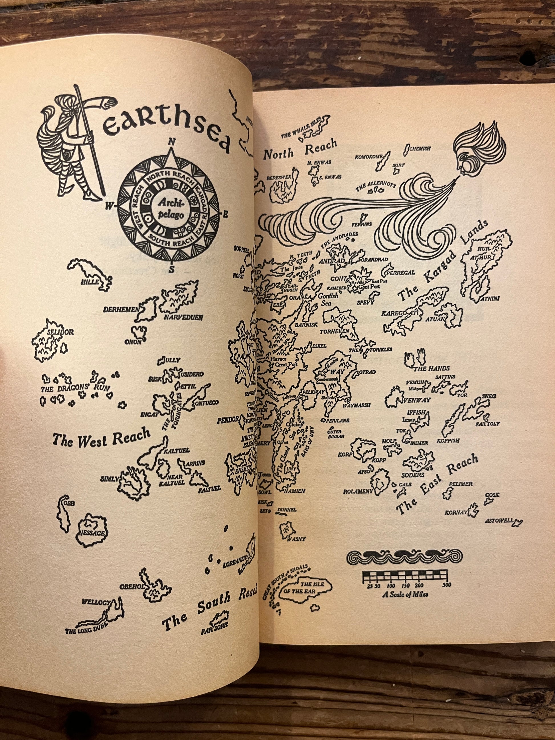 Open book showing a map of Earthsea, the book is titled 'a Wizard of Earthsea' by Ursula K. Le Guin on a wooden surface 