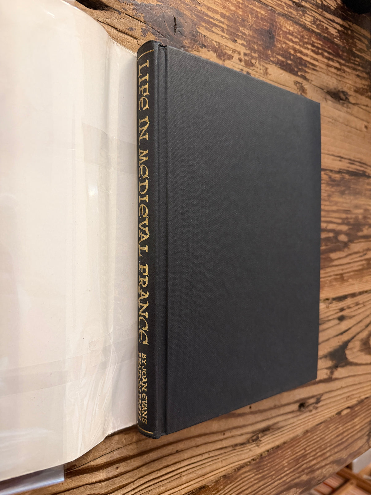 The front cover and spine without dust jacket of a book titled 'Life in Medieval France' by Joan Evans, on a wooden surface