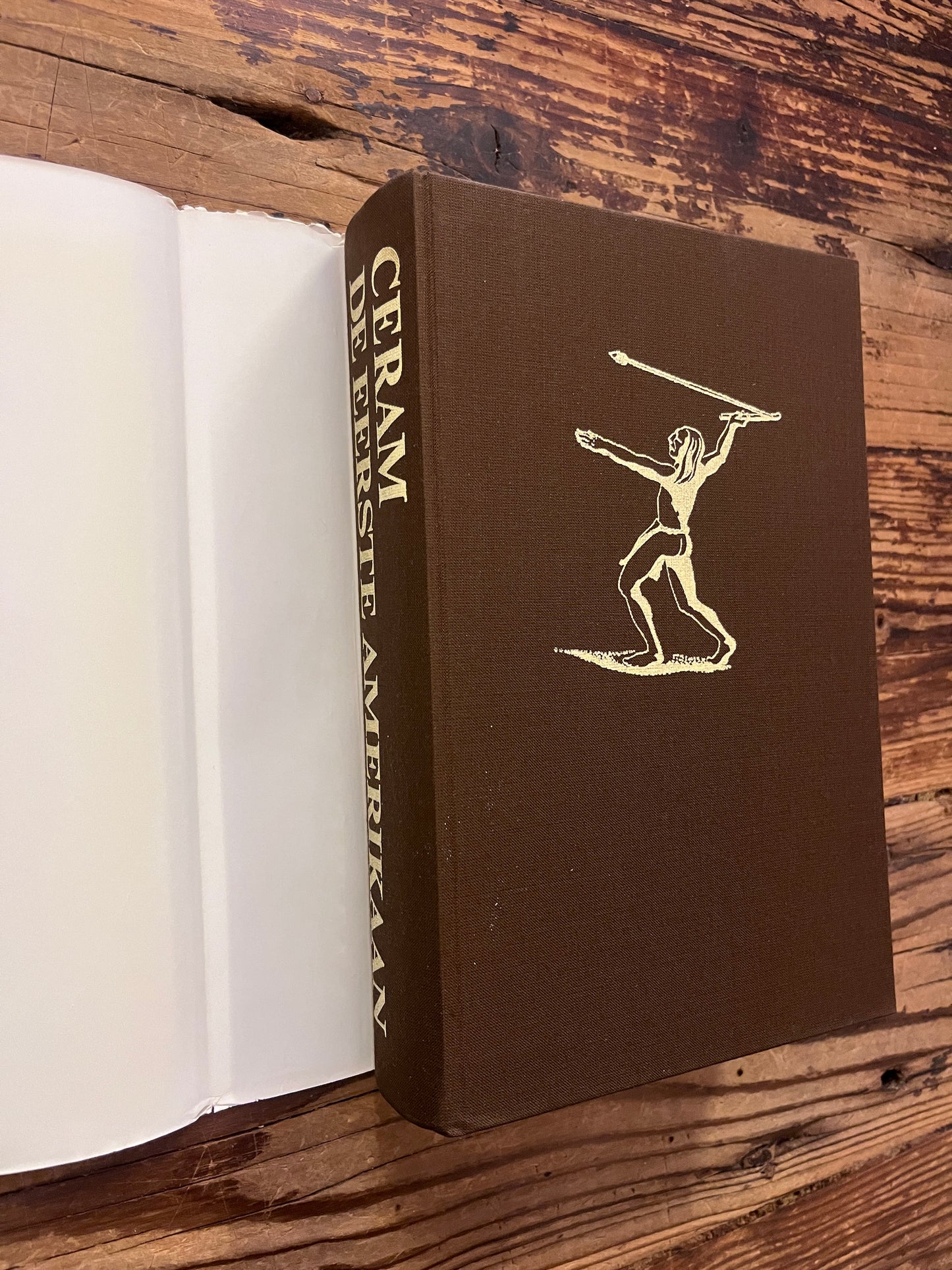 Voorkant en rug zonder stof omslag van een hardcover boek met de titel 'De Eerste Amerikaan' by C.W. Ceram op een houten achtergrond 