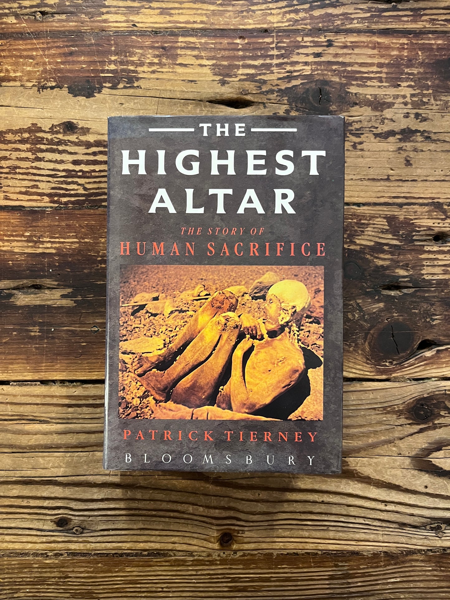 Front cover of a book with the title 'The Highest Altar: The Story of Human Sacrifice' by Patrick Tierney is displayed on a wooden surface
