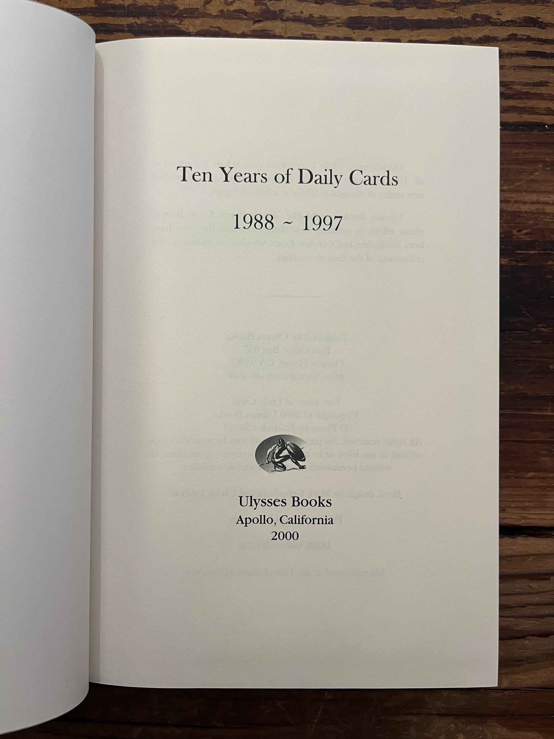 Open book with the title 'Ten Years of Daily Cards' by Robert Burton on a wooden surface