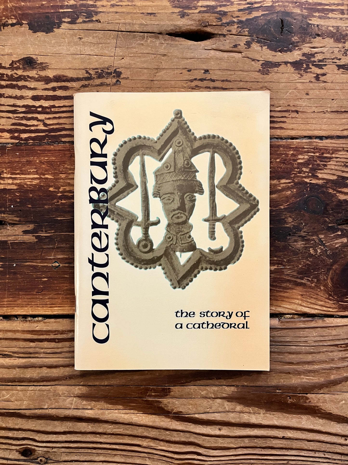 Front cover of a book titled 'Canterbury: the story of a cathedral' with a beige cover, featuring a large ornate cathedral emblem in the center and vertical 'Canterbury' text on the left, photographed on a rustic wooden surface.