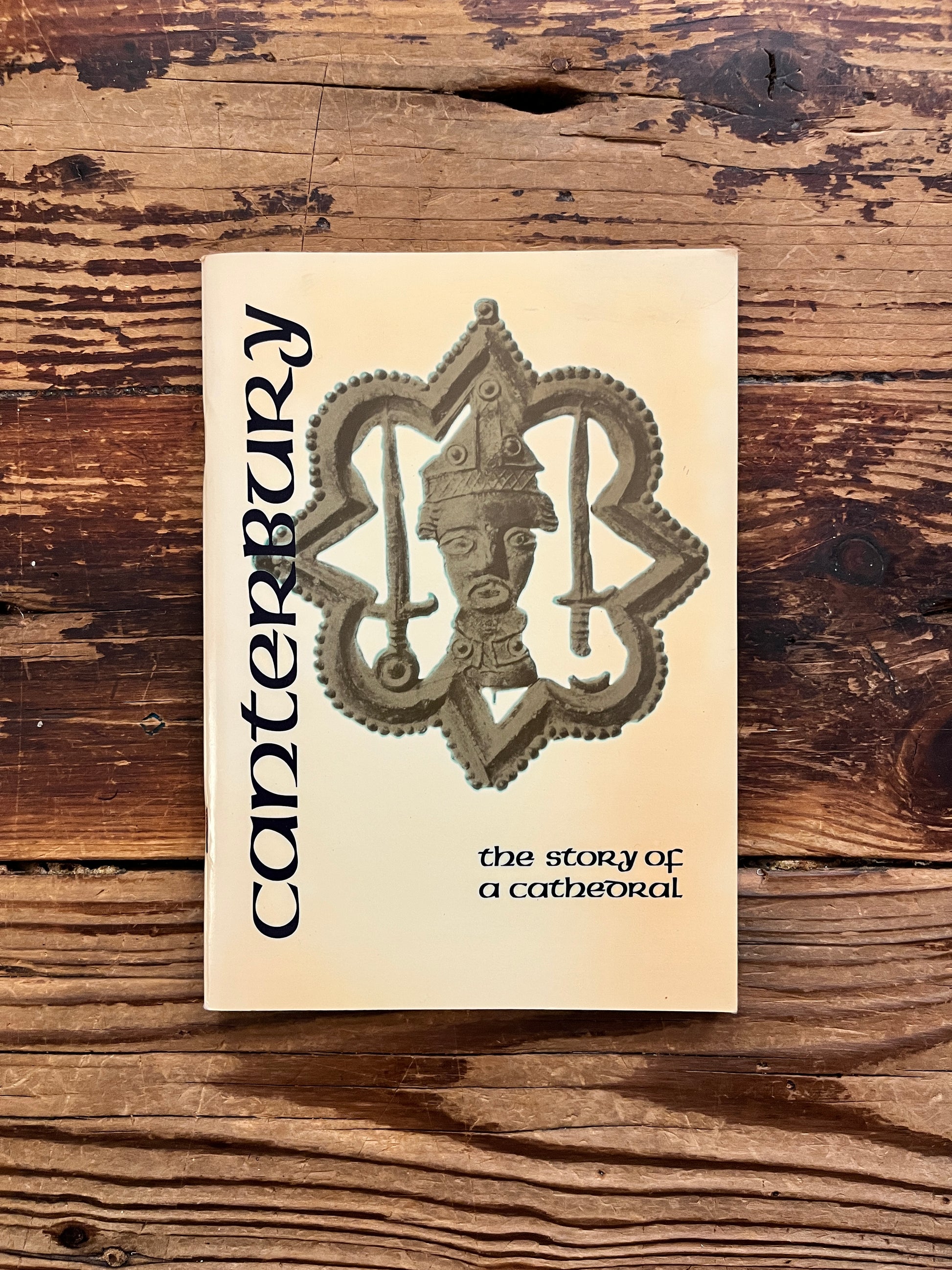 Front cover of a book titled 'Canterbury: the story of a cathedral' with a beige cover, featuring a large ornate cathedral emblem in the center and vertical 'Canterbury' text on the left, photographed on a rustic wooden surface.