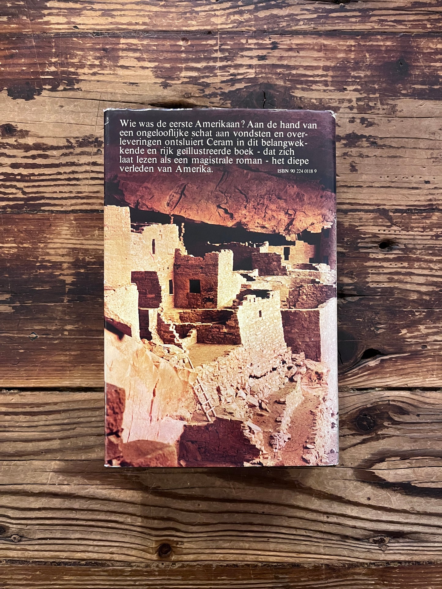 Achterkant van een hardcover boek met de titel 'De Eerste Amerikaan' by C.W. Ceram op een houten achtergrond 