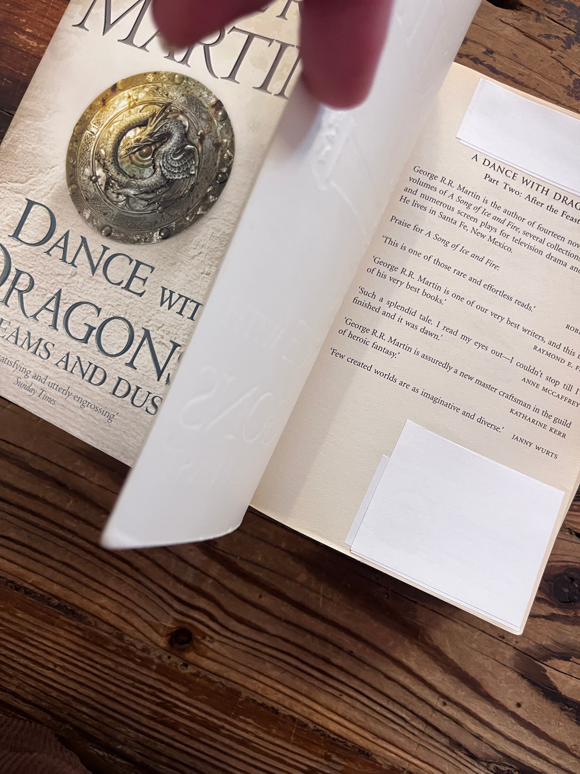 Corner with white sticker of the first page of Two books titled 'A Dance with Dragons' by George R.R. Martin on a wooden surface.