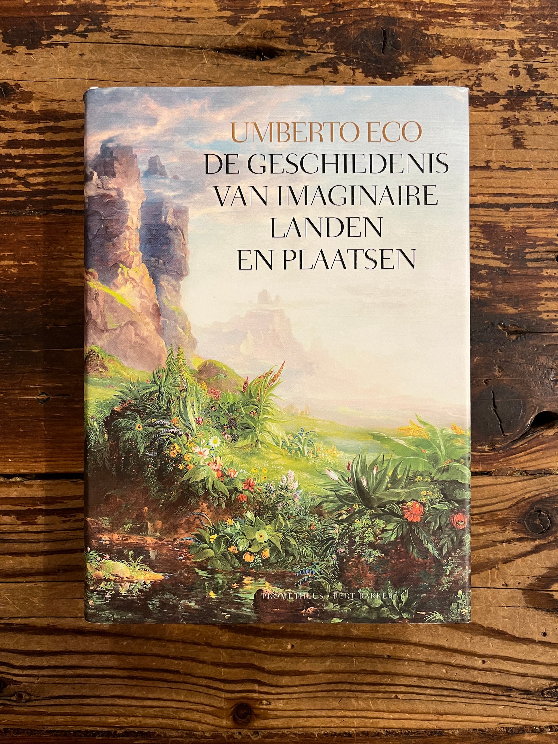 Voorkant van boek met de titel 'De Geschiedenis Van Imaginaire Landen En Plaatsen' door Umberto Eco op een houten achtergrond 