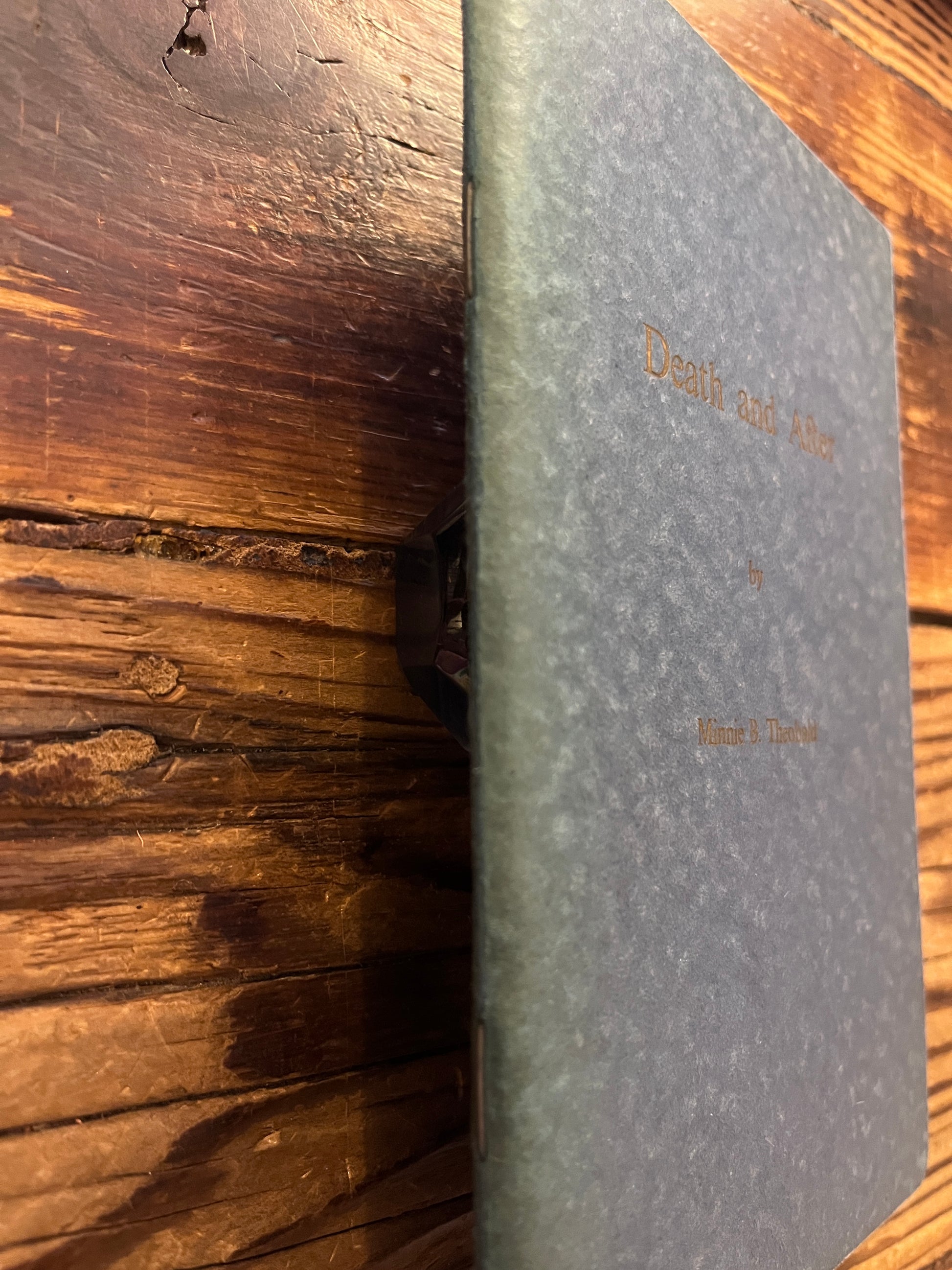 Spine of the stapled booklet showing the staples from the outside. Booklet has the title 'Death and After' by Minnie B. Theobald on a wooden surface 