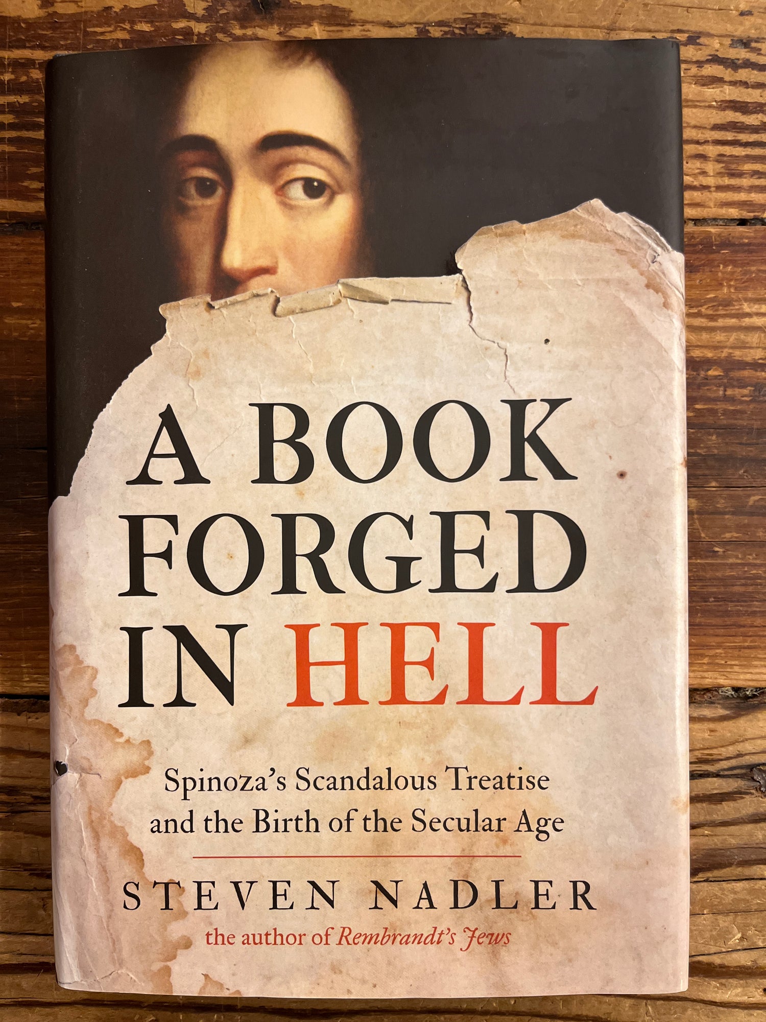front cover of book titled 'A Book Forged in Hell' by Steven Nadler on a wooden surface