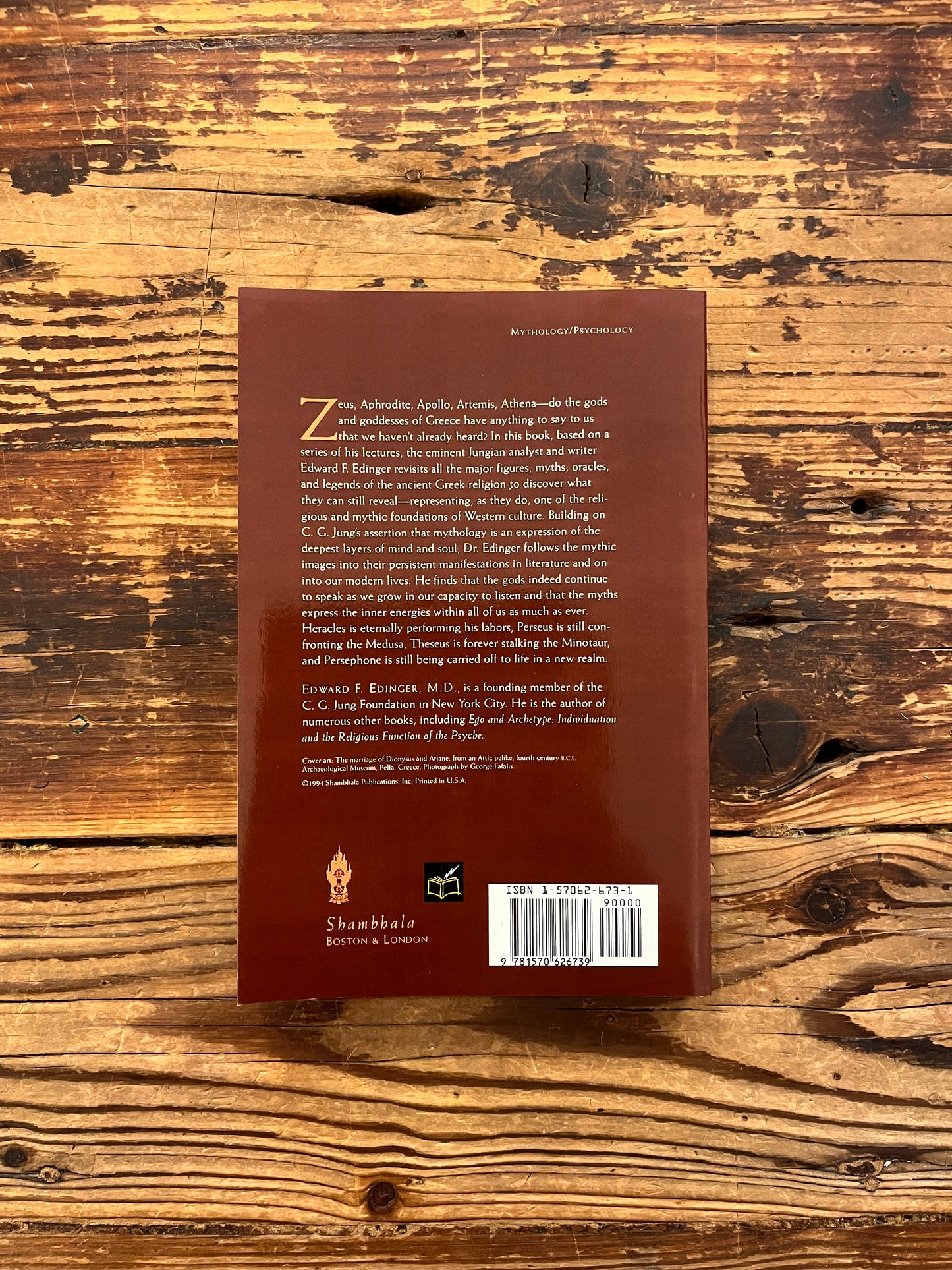 Back cover of book titled 'The Eternal Drama: Greek Mythology' by Edward T. Edinger lying on a wooden surface