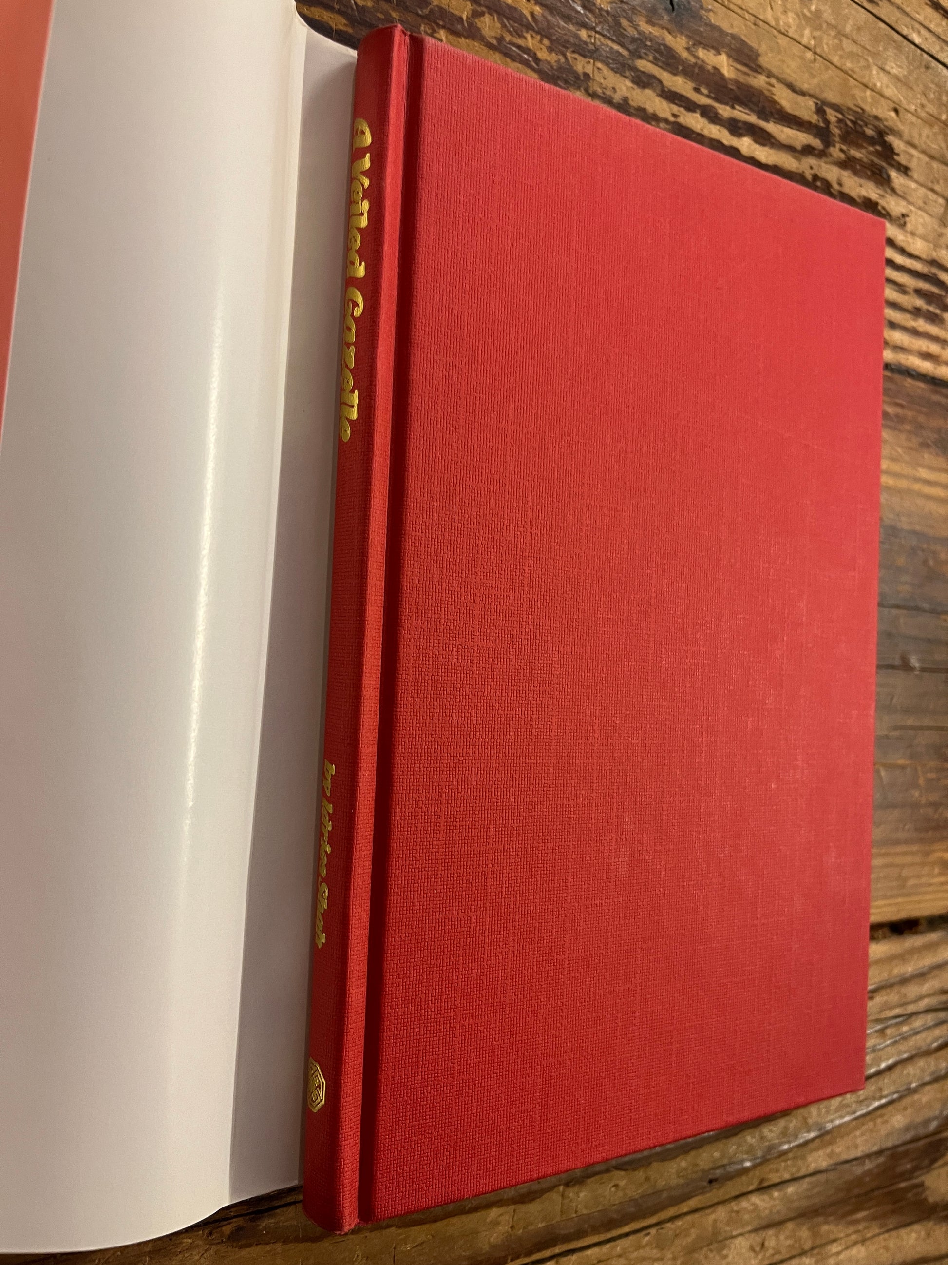 Red book titled 'A Veiled Gazette' by Idries Shah with gold text on a wooden surface