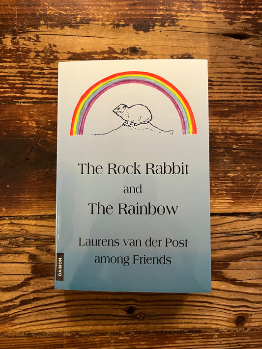 Book cover of 'The Rock Rabbit and The Rainbow' by Laurens van der Post on a wooden surface