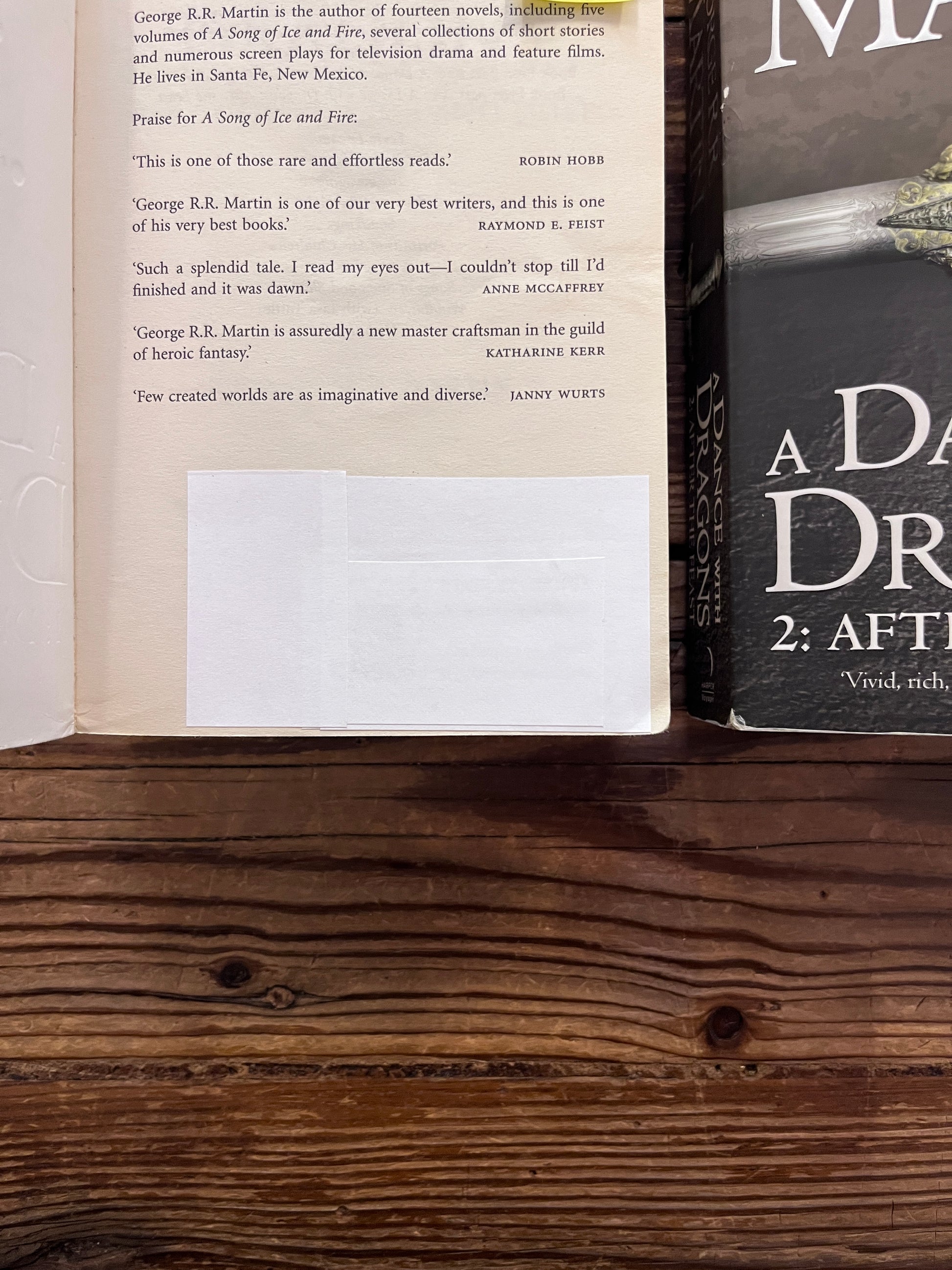 Corner with white sticker of the first page of Two books titled 'A Dance with Dragons' by George R.R. Martin on a wooden surface.