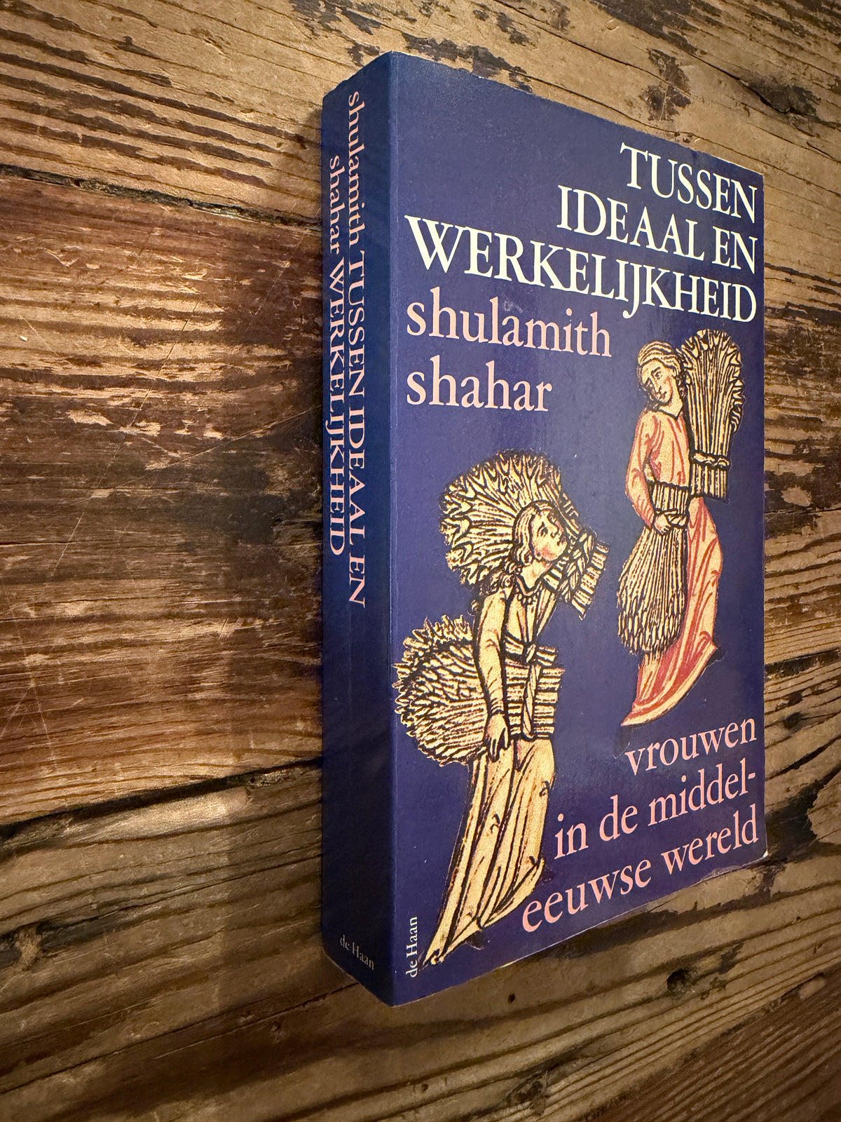 Voorkant en rug van boek met de titel 'Tussen ideaal en werkelijkheid, vrouwen in de middeleeuwse wereld' door Shulamith Shahar op een houten achtegrond 